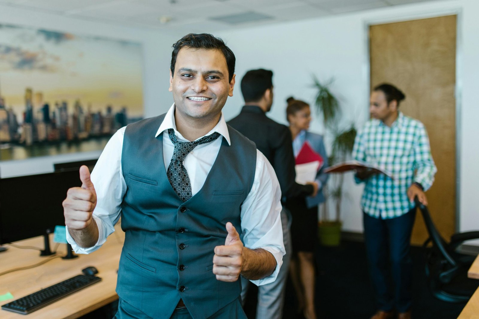 Smiling businessman showing thumbs up in a lively office environment with colleagues in the background.
